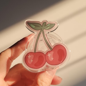 Peut inclure: Un charm en acrylique transparent en forme de cerises, avec deux cerises rouges et des feuilles vertes. Le charme est présenté sur un fond clair, mettant en valeur les détails du design et l'effet de la lumière du soleil.