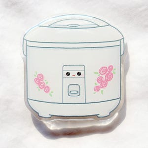 Pode incluir: Um adesivo branco de panela de arroz em estilo cartoon com contorno azul claro. O adesivo apresenta detalhes de rosas cor-de-rosa e um rosto sorridente. O design é simples e fofo, ideal para decorar laptops, cadernos ou outros itens.