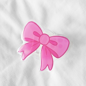 May include: A pink bow-shaped sticker with a watercolour effect. The bow has two loops on each side and a central knot. The sticker is on a clear, circular base, set against a white, textured background.