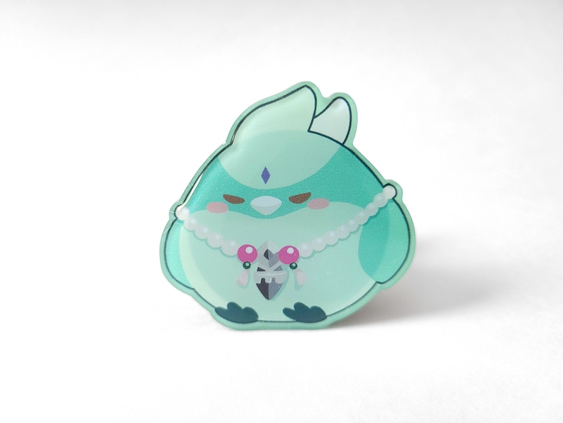 May include: A green and white cartoon bird pin with a pearl necklace and a pink gem pendant. The bird has a small, white beak and a blue gem on its forehead.