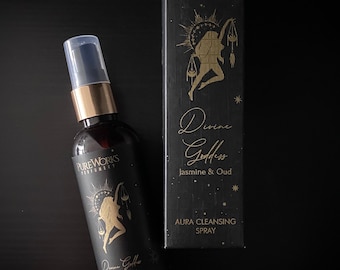 Può includere: Un flacone nero con un ugello spray dorato e un'etichetta. L'etichetta presenta un'illustrazione dorata di una donna che tiene una bilancia e il testo "Divine Goddess Jasmine & Oud Aura Cleansing Spray".