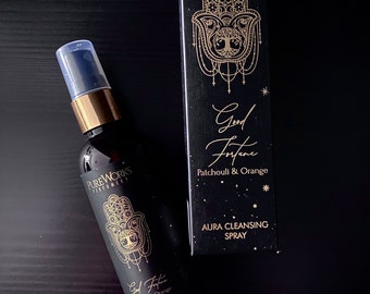 Può includere: Una bottiglia nera con un'etichetta dorata che dice "Pure Works Perfumery" e "Good Fortune Patchouli & Orange Aura Cleansing Spray". La bottiglia è su una superficie nera. L'etichetta ha un design dorato a mano di Hamsa.