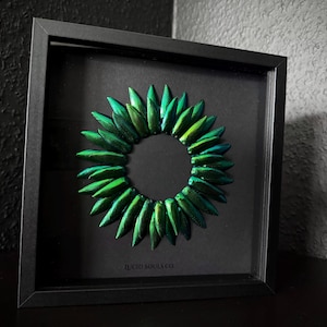 Könnte beinhalten: Gerahmtes Kunstwerk mit einem kreisförmigen Arrangement aus schillernden grünen Käferflügeln. Die Flügel sind um einen zentralen Leerraum angeordnet und erzeugen einen auffälligen visuellen Effekt. Der Rahmen ist schwarz, der Hintergrund dunkel. Der Text "LUCID SOULS CO" steht unten.