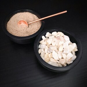 May include: Two black bowls, one filled with white gravel and the other with brown sand. A small copper spoon rests on top of the sand.