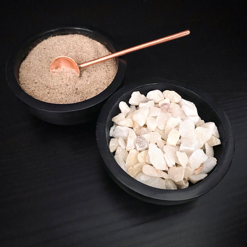 May include: Two black bowls, one filled with white gravel and the other with brown sand. A small copper spoon rests on top of the sand.