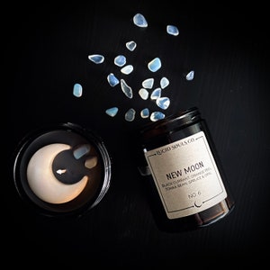 Könnte beinhalten: Ein schwarzes Kerzengefäß mit einem weißen Halbmond-Design und dem Text "LUCID SOULS CO. NEW MOON BLACK CURRANT, ORANGE PEEL, TONKA BEAN, SPRUCE & ORL NO. 6" auf dem Etikett. Das Gefäß ist von kleinen, hellblauen, schillernden Steinen umgeben.
