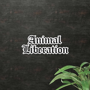 May include: A white sticker with the words "Animal Liberation" in a gothic font. The sticker is set against a dark wood-grain background, with a few green leaves in the lower right corner.