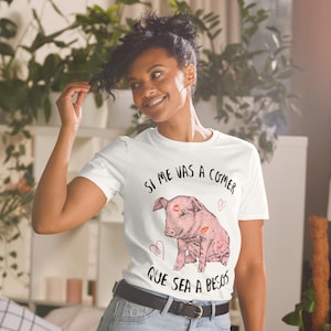 May include: White t-shirt featuring a pink pig illustration with lipstick kisses and the Spanish text "Si me vas a comer que sea a besos." The design includes hearts. The shirt is short-sleeved.