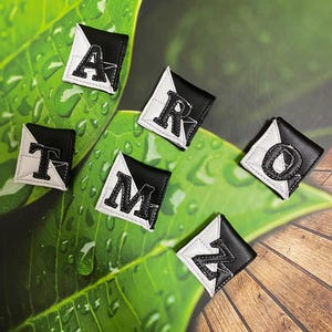 May include: Black and white letter patches arranged on a green leaf surface. Each patch features a different letter: A, R, T, M, O, and Z. The patches have a black background with a white border and a white letter.