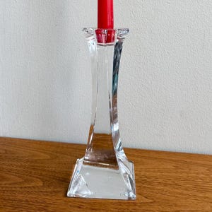 Puede incluir: Un candelero de vidrio transparente y facetado con una vela cónica roja. La vela es alta y delgada, y el soporte tiene una base cuadrada y una parte superior acampanada. El candelero está sobre una superficie de madera.