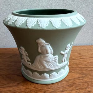 May include: A sage green ceramic planter featuring white relief ornamentation. The design includes a central seated figure flanked by two cherubs, with decorative leaf-patterned borders encircling both the upper rim and the base of the vessel.
