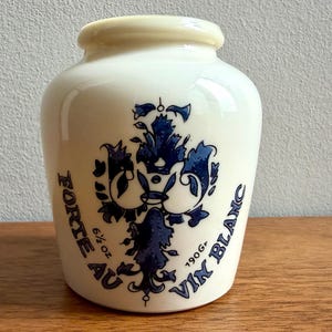 May include: A cream-colored ceramic jar with a smooth, rounded body and a short neck. The front features a dark blue floral emblem and the printed text "FORTE AU VIN BLANC" along with the specific weight measurements of 6 1/2 oz and 190 gr.