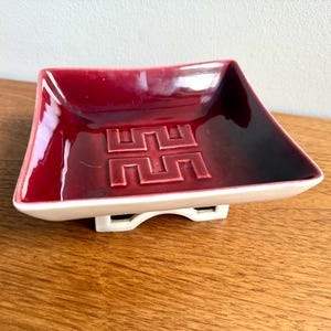 May include: A square, burgundy-colored ceramic dish with a geometric design in the center. The dish has a glossy finish and sits on a white base. The dish is on a wooden surface.