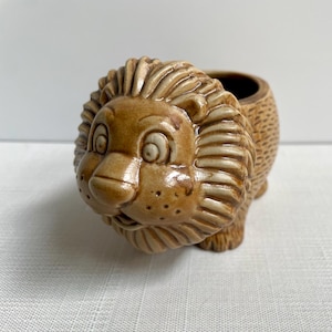 May include: A ceramic lion-shaped container with a brown and tan glaze. The lion's face is detailed with a wide-eyed expression and a playful smile. The container is open and appears to be a trinket box.