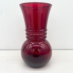 May include: A ruby red glass vase with a bulbous base and a flared top. The vase has a series of horizontal ridges around the middle. The glass is transparent, allowing light to pass through and create reflections. The vase is likely used for displaying flowers or as a decorative item.