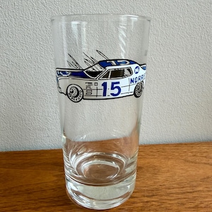 May include: A clear glass tumbler featuring a vintage race car design. The car is white and blue with the number 15 prominently displayed. The glass has a classic, straight-sided shape, perfect for serving beverages.