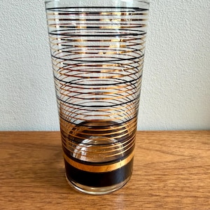 May include: A clear glass tumbler with a gold and black striped design. The stripes are horizontal and wrap around the glass. The bottom of the glass has a thick black band with a gold stripe. The glass is on a wooden surface.