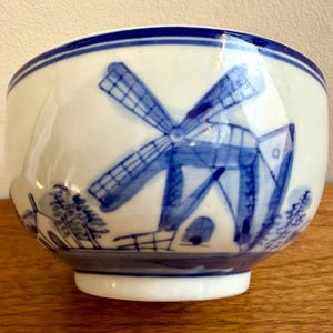 May include: A white ceramic bowl with a blue windmill design. The bowl has a rounded shape with a blue rim and a detailed illustration of a windmill and landscape in blue. The bowl is on a wooden surface.