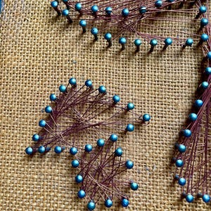 Pode incluir: Um close-up de uma pe&ccedil;a de arte de cordas com contas azuis e fio de cobre. A arte est&aacute; em um fundo de juta.