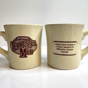 May include: Two white ceramic mugs with brown text. One mug says "Jack Daniel's Tennessee Mud" and the other mug says "1 Part Jack Daniel's 1 Part Amaretto Whipped Cream Coffee".