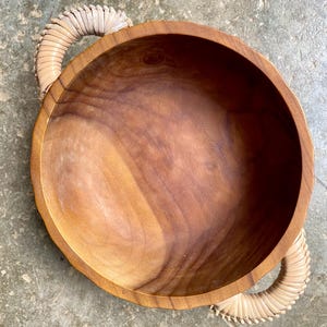 May include: A large, round wooden bowl with two woven handles. The bowl is made of light brown wood and has a natural finish. The handles are made of a light beige material and are attached to the sides of the bowl.