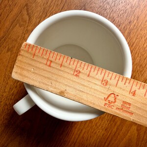 May include: A white ceramic teacup with a handle, viewed from above, with a wooden ruler laid across the cup's opening. The ruler is marked in inches, showing the cup's diameter. The cup sits on a wooden surface.