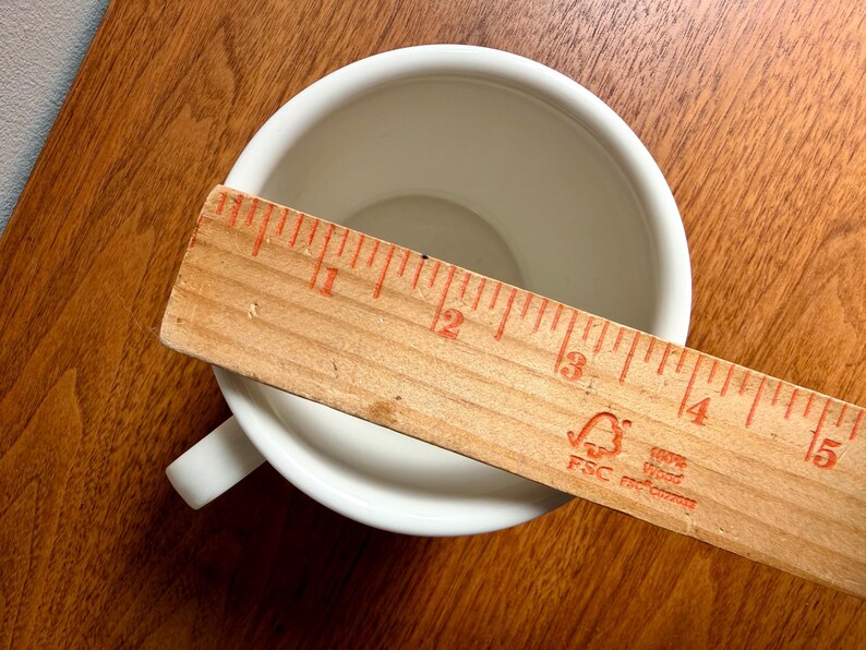 May include: A white ceramic teacup with a handle, viewed from above, with a wooden ruler laid across the cup's opening. The ruler is marked in inches, showing the cup's diameter. The cup sits on a wooden surface.