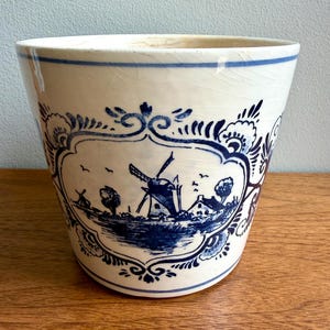 May include: Cream-colored ceramic planter with a blue windmill and landscape design. The planter features a detailed illustration of a Dutch windmill, trees, and a house. The rim has a blue stripe and the design is reminiscent of Delftware.
