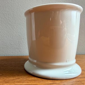 May include: A white ceramic shaving mug with a rounded base and a flared top. The mug has a smooth, glossy finish and is set on a wooden surface. The mug is designed for holding shaving cream or soap.