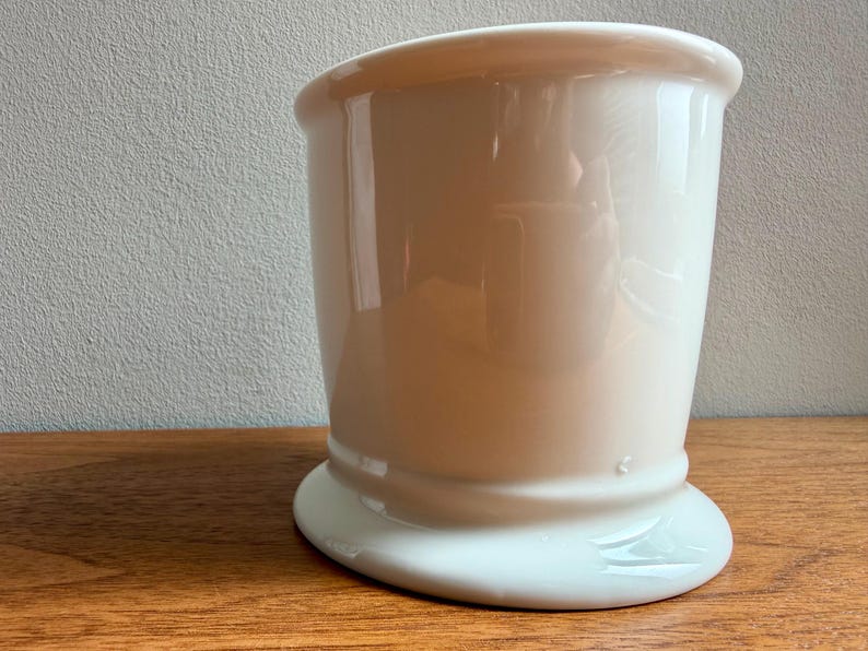 May include: A white ceramic shaving mug with a rounded base and a flared top. The mug has a smooth, glossy finish and is set on a wooden surface. The mug is designed for holding shaving cream or soap.