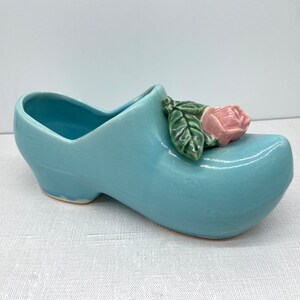 May include: A light blue ceramic planter shaped like a clog with a pink rose on top.