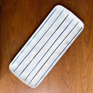 May include: A rectangular white serving tray with rounded corners, featuring a series of thin, parallel dark blue stripes running diagonally across the surface. The tray has a smooth, glossy finish and a clean, simple design for serving food.