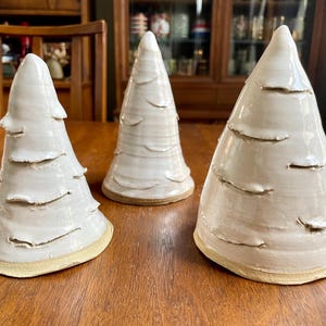 May include: Three ceramic Christmas tree decorations. Each tree is white with horizontal textured bands and a pointed top. The trees are arranged on a wooden table. The base of each tree is a light tan color.
