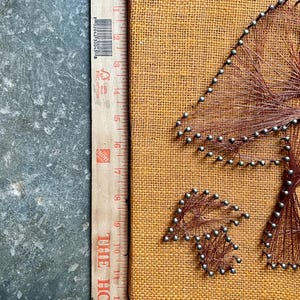 Pode incluir: Uma tela de juta marrom com um design de arte de arame de cobre e pregos que representa uma &aacute;rvore com folhas. A arte est&aacute; montada em uma moldura de madeira.
