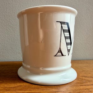 May include: A white ceramic shaving mug with a flared top and a wide base. The letter "M" is prominently displayed in black, featuring a striped design. The mug sits on a wooden surface.