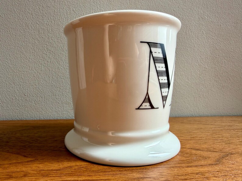 May include: A white ceramic shaving mug with a flared top and a wide base. The letter "M" is prominently displayed in black, featuring a striped design. The mug sits on a wooden surface.