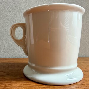 May include: A cream-colored ceramic shaving mug with a handle. The mug has a wide, curved rim and a flared base. The handle is a loop shape. The mug is sitting on a wooden surface.