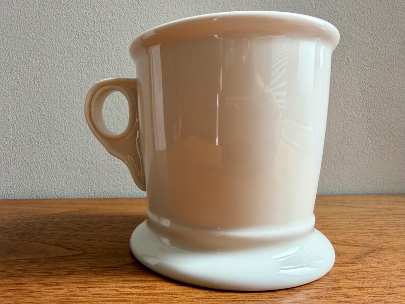 May include: A cream-colored ceramic shaving mug with a handle. The mug has a wide, curved rim and a flared base. The handle is a loop shape. The mug is sitting on a wooden surface.