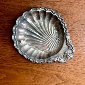 May include: A silver-toned metal dish shaped like a scallop shell. The dish features a scalloped edge and a decorative embossed floral pattern on the handle area. The interior surface has radiating fluted lines extending from the base to the rim.