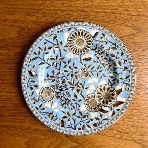 May include: A round ceramic plate featuring a light blue background with a detailed pattern of dark brown floral motifs, including large stylized sunflowers, intricate leafy branches, and small birds nestled among the foliage, finished with a geometric border.
