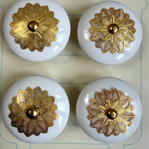 May include: Set of four white ceramic drawer knobs with gold filigree floral designs. Each knob features a central gold accent. The knobs are from Cynthia Rowley, New York.