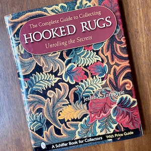 May include: A book titled "The Complete Guide to Collecting Hooked Rugs" by Jessie A. Turbayne. The cover features a floral pattern in shades of red, blue, yellow, and green against a dark background. The book is a Schiffer Book for Collectors.