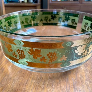 May include: A clear glass serving bowl with a wide rim. The bowl is decorated with a gold and green leaf and grape design. The bowl is sitting on a wooden surface.