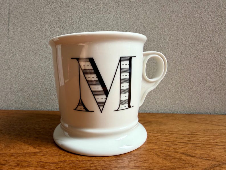 May include: White ceramic mug with a large black letter "M" on the front. The letter is decorated with horizontal stripes and dashes. The mug has a rounded base and a handle. The mug is sitting on a wooden surface.