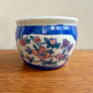 May include: A small, round ceramic pot with a blue and white design. The pot features a band of blue around the top and bottom, with a white section in the middle. The white section is decorated with pink flowers and green leaves.