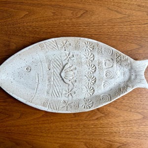 May include: A ceramic fish-shaped serving platter with a speckled, off-white finish. The platter features embossed floral and geometric patterns. The fish measures approximately 15 inches long.