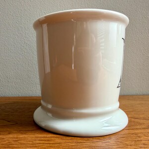 May include: A cream-colored ceramic shaving mug with a wide base and a flared top. The mug has a black graphic on the side. The mug is sitting on a wooden surface.