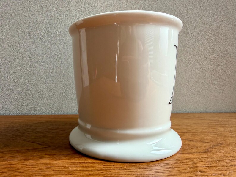 May include: A cream-colored ceramic shaving mug with a wide base and a flared top. The mug has a black graphic on the side. The mug is sitting on a wooden surface.