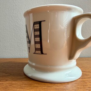 May include: Cream-colored ceramic mug with a wide base and a large handle. The letter "M" is prominently displayed in black, with a striped design. The mug is set on a wooden surface.