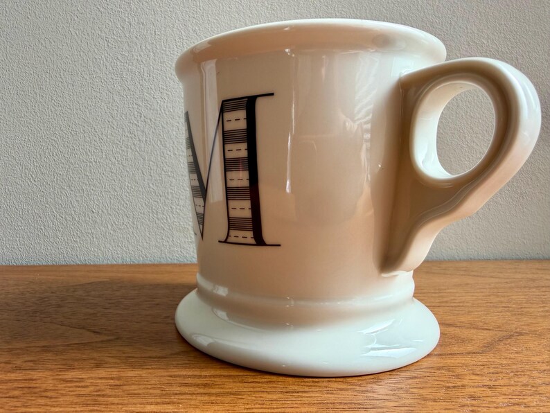 May include: Cream-colored ceramic mug with a wide base and a large handle. The letter "M" is prominently displayed in black, with a striped design. The mug is set on a wooden surface.